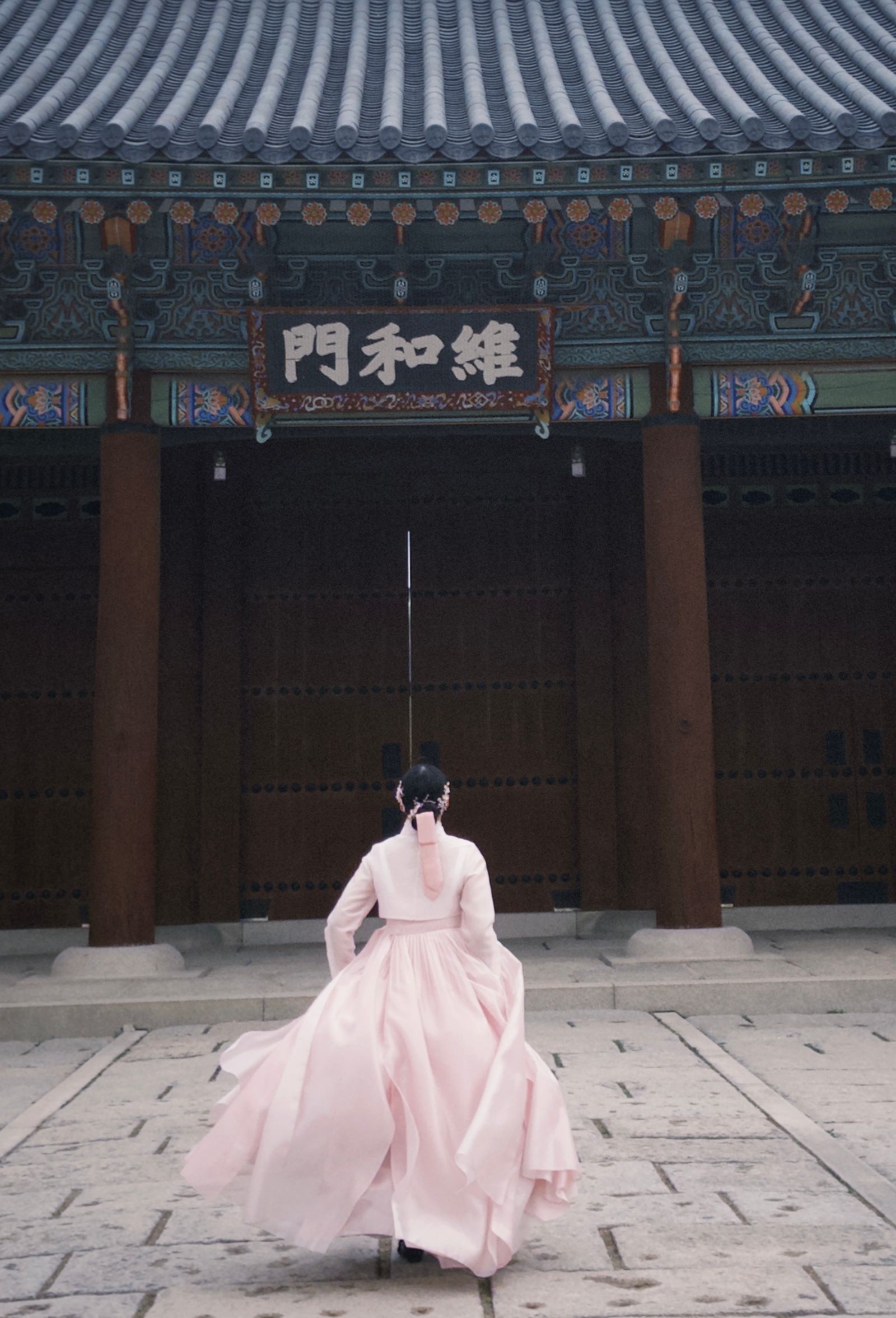 Une femme en hanbok rose, de dos, devant la porte d’un palais de Séoul